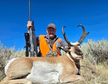 A hunter, holding a rifle, poses triumphantly with a pronghorn antelope in the sagebrush of Wyoming, set against a clear, vivid blue sky.